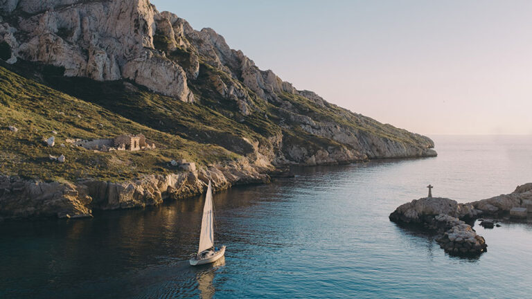 A sail boat sailing through the blue ocean on a summers morning with cliffs and land in the distance.