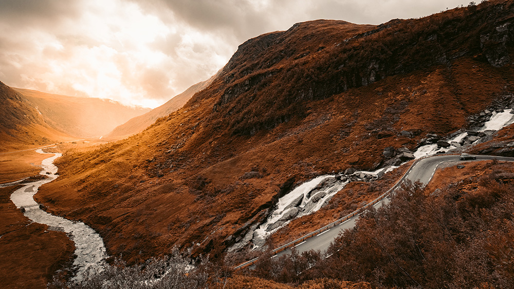 A deep orange cliff with a winding road going up it with a lone car on it. There is a winding flow of water going across the cliff aswell.
