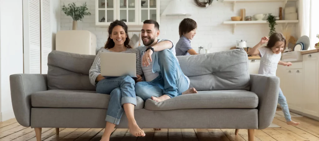 A family in a living room. A man and woman are sat on the sofa smiling on a laptop and their young son and daughter are running around the sofa smiling.