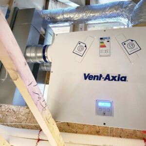 A close up of a ventilation system with a blue logo saying "Vent-Axia" on it aswell as a digital display and a energy consumption sticker on it, surrounding by metal pipes.