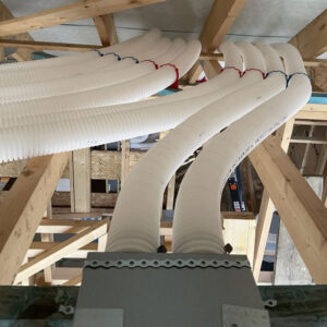 A close up of an attic full of plastic white pipes and exposed wooden beams and a blue and red ventilation system with metal ducts sitting on the beams.