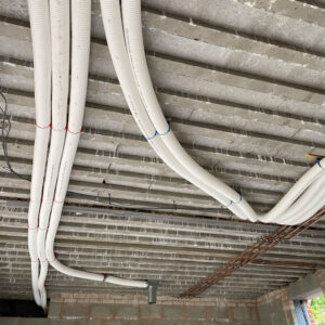 A series of white plastic pipes, bronze pipes and wires running across a concrete ceiling in a building under construction.