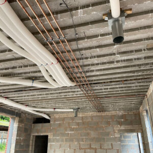 A series of white plastic pipes, bronze pipes and wires running across a concrete ceiling in a building under construction.