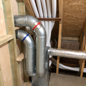 A close up of metal pipes and white plastic pipes inside a wooden attic.