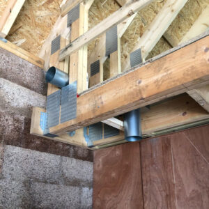 A close-up photo of a weathered wooden frame of a house under construction. The frame consists of vertical and horizontal beams connected with metal brackets. Diagonal beams are visible in the background. There is a metal pipe with a hole in it attached to the frame.