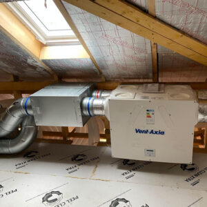 A ventilation system with a blue logo saying "Vent-Axia" on it aswell as a digital display and a energy consumption sticker on it, surrounding by metal pipes. In an attic with a window above it.