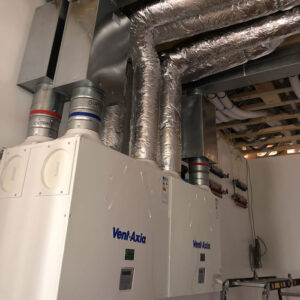 A close-up view of two ventilation systems with energy consumption stickers on them. The systems consists of exposed metal pipes with the brand name “Vent Axia” printed on the unit in blue.