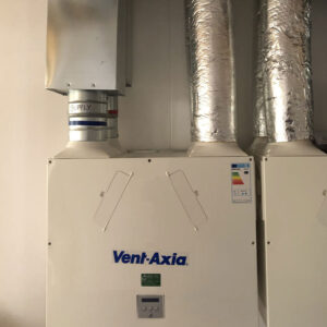 A close-up view of a ventilation system with an energy consumption sticker on it. The system consists of exposed metal pipes with the brand name “Vent Axia” printed on the unit in blue.