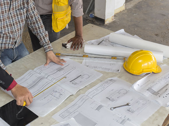 Two construction workers leaning on a table looking at floor plans. There is a yellow helmet and several tools on the desk.