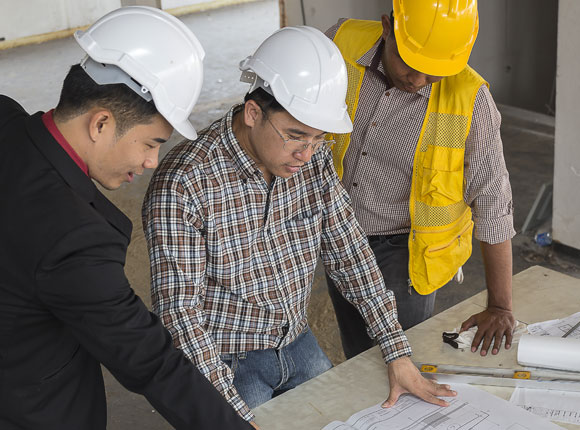Three construction workers with helmets on looking together at a floor plan on a desk.