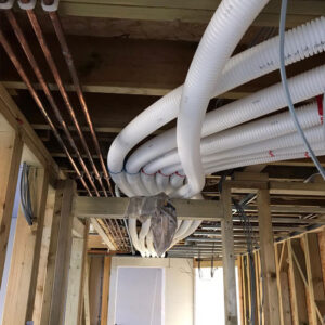 A bunch of exposed pipes hang from the ceiling of a building under construction. The pipes are various sizes and colours, including metal and insulated white pipes.