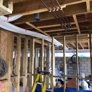 A close-up view of a building under construction. Exposed pipes of various sizes and materials, including metal and insulated white pipes, hang from the unfinished ceiling.