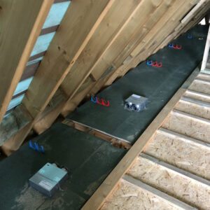 An attic floor with a ventilation system installed on it. The ventilation system consists of metal ducts with insulation wrapped around them. The ducts connect to a box in the centre of the image. There are also wooden beams visible.