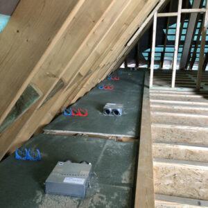 An attic floor with a ventilation system installed on it. The ventilation system consists of metal ducts with insulation wrapped around them. The ducts connect to a box in the centre of the image. There are also wooden beams visible.