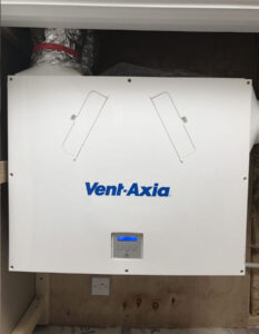 A close-up view of a ventilation system with a digital display in a building under construction. The system consists of exposed metal pipes with the brand name “Vent Axia” printed on them.