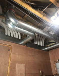 A bunch of exposed pipes hang from the ceiling of a building under construction. The pipes are various sizes and colours, including metal and insulated white pipes.