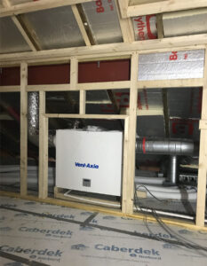 A close-up view of a ventilation system in a building under construction. The system consists of exposed metal pipes with the brand name “Vent Axia” printed on them.