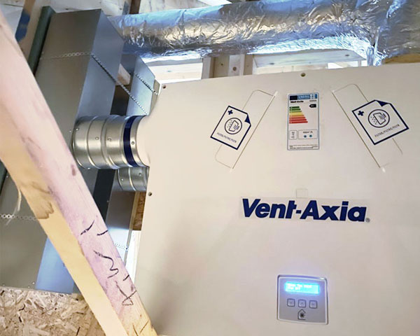 A close-up view of a ventilation system with a digital display in a building under construction. The system consists of exposed metal pipes with the brand name “Vent Axia” printed on them and an energy consumption sticker.
